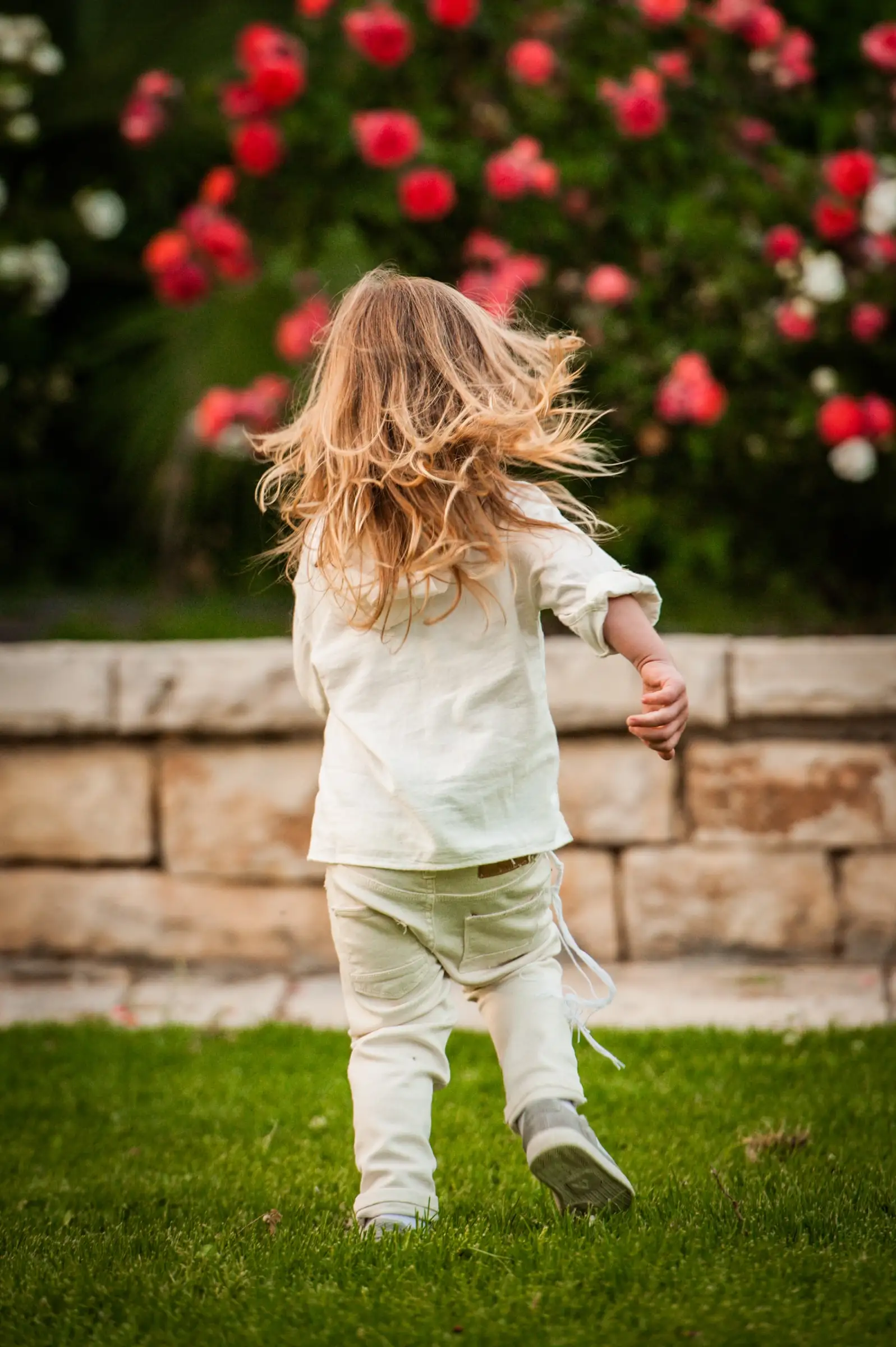 A boy running across grass, his long hair flying, against a Jerusalem stone wall