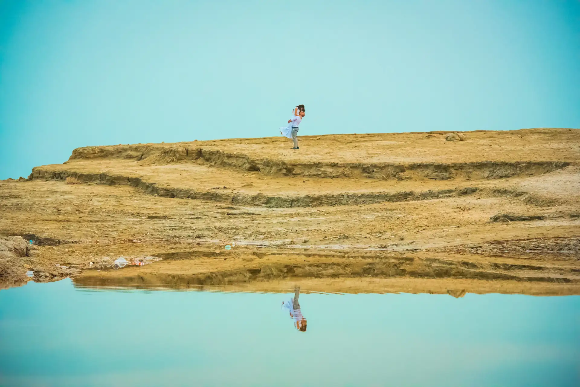 A couple mid-lift on a desert ridge, reflected in a salt pool